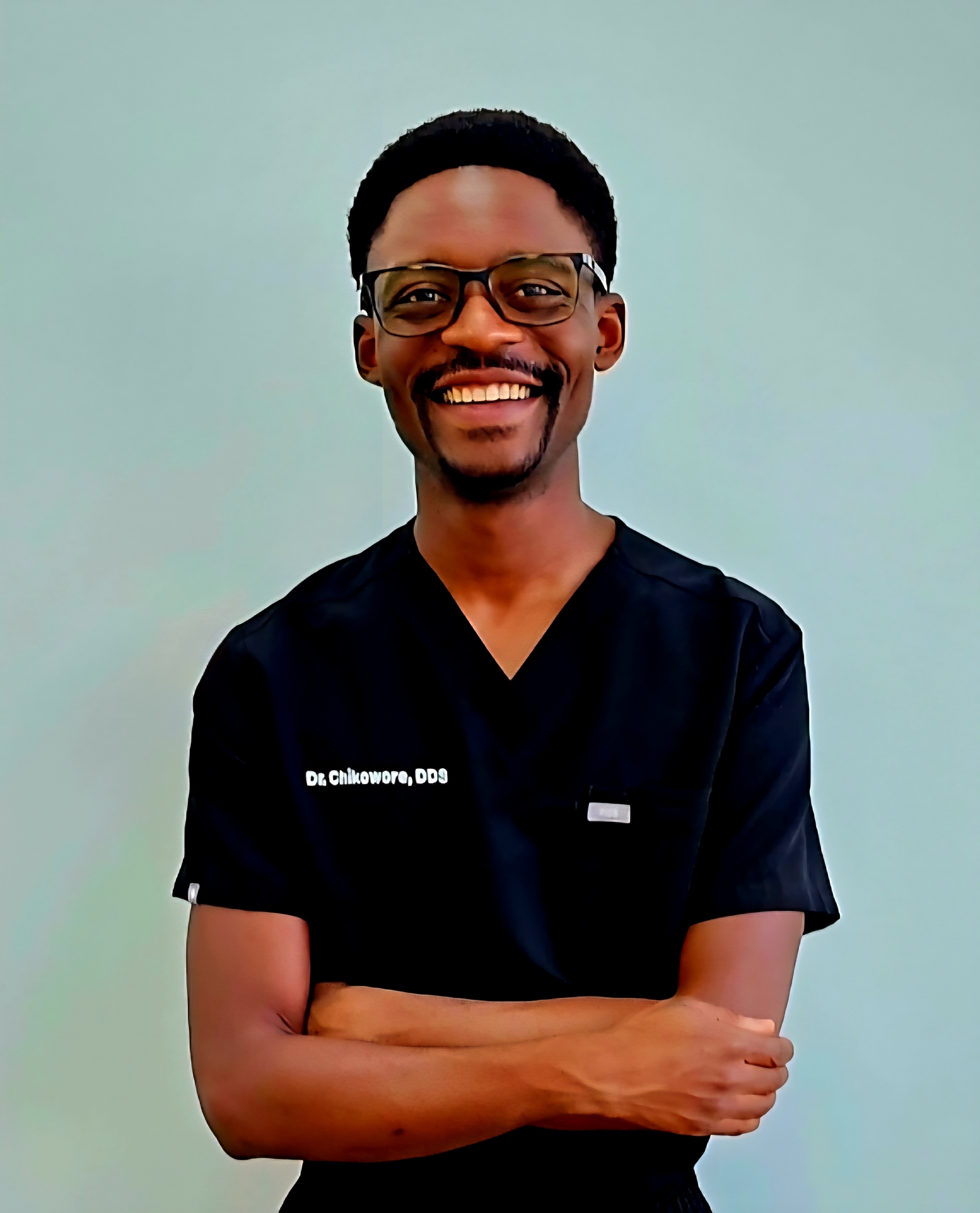 Dr. Tapuwa Chikowore, General Dentist - Professional headshot showing him in navy blue dental scrubs with 'Dr Chikowore, DDS' embroidered on the chest, wearing glasses and smiling warmly with arms crossed